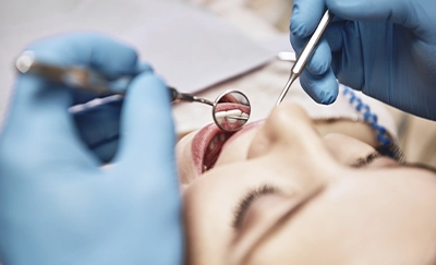 Dentist in blue gloves using a dental mirror and tool to examine a patient’s open mouth during a dental procedure.