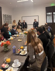 Business meeting in a conference room with a presenter at the head of a long table as colleagues listen over coffee and pastries.