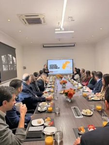 Professionals seated around a long conference table with snacks and orange juice, listening to a presenter at the front with a screen display.