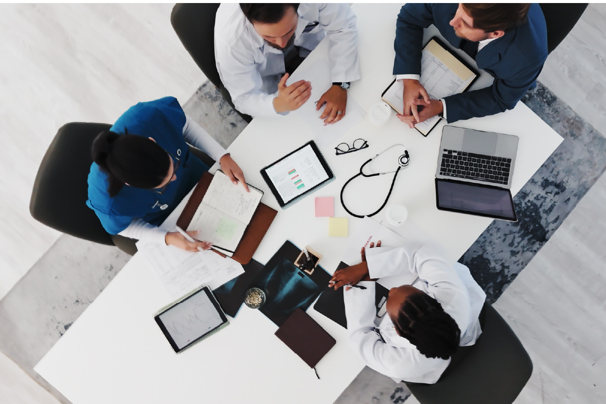 Overhead view of a diverse medical team around a white conference table, reviewing tablets, notebooks, stethoscope, laptop, and sticky notes as they collaborate.
