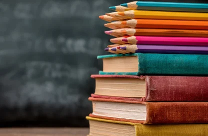 Stack of colorful pencils on top of stacked hardcover books in a classroom setting with a chalkboard in the background.
